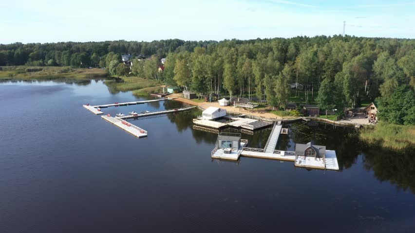 Aerial view of a peaceful lakeside marina with floating docks, boats, forest background, and a recreational area surrounded by nature on a sunny day.
