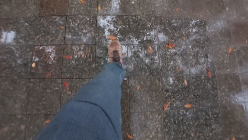 Time lapse, POV: person's legs fast going in rain on damp granite paving slabs. Point of view of men's feet are dressed in jeans and boots briskly walking along wet sidewalk covered in autumn leaves
