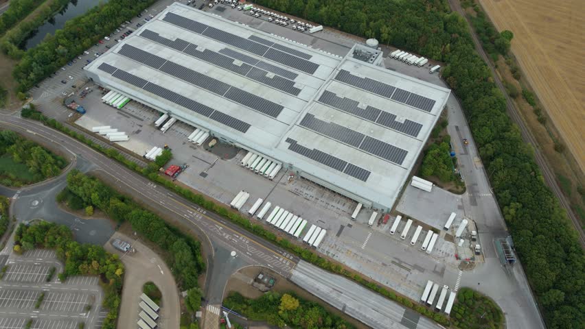 Aerial view of a large building with solar panels and many trucks in the loading bay surrounded by green trees and roads, Bedford, England, United Kingdom.