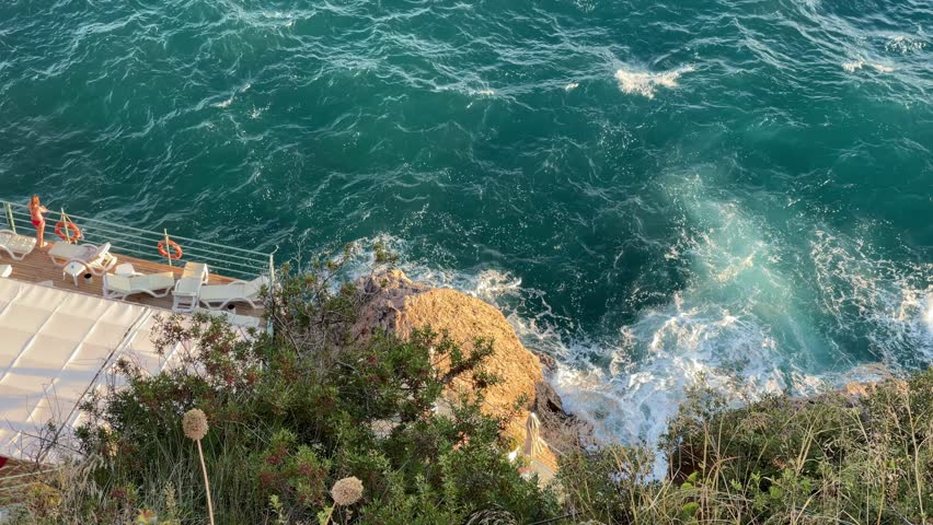 Scenic coastal pathway while large waves crash against the rocks below. The bright sun highlights the beautiful turquoise water of Mediterranean Sea