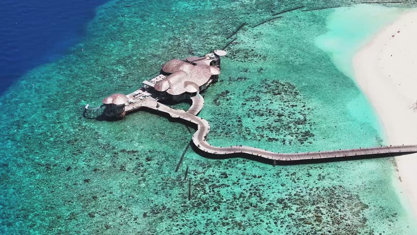 Aerial view of a thatched roof resort connected by a winding wooden walkway over the turquoise waters, contrasting with the white sandy beach, Eydhafushi, Baa Atoll, Maldives.