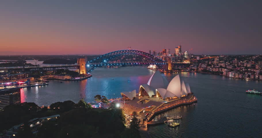 Australia, Sydney: Sydney Harbour at sunset, illuminated Sydney Opera House and Harbour Bridge against vibrant sky. Breathtaking aerial view of modern city skyline at night. Drone flight footage