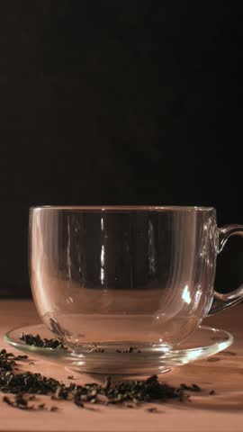 Hot water is poured into a transparent glass cup with green tea leaves, steam rising, capturing freshness and morning tranquility.