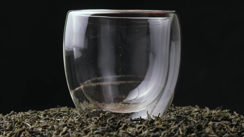 Green tea is poured into a slowly rotating glass, swirling golden infusion among tea leaves in macro close-up on black background.