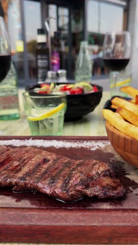Vertical video. A skirt steak with French fries on the table with a salad against the backdrop of glasses of red wine, dining out concept. High quality 4k footage