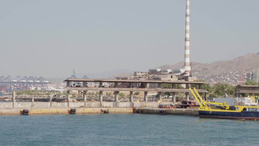 A concrete pier with an abandoned control tower extends into the water. Behind it, cranes, smokestacks, and dense urban buildings rise against a mountainous backdrop. 