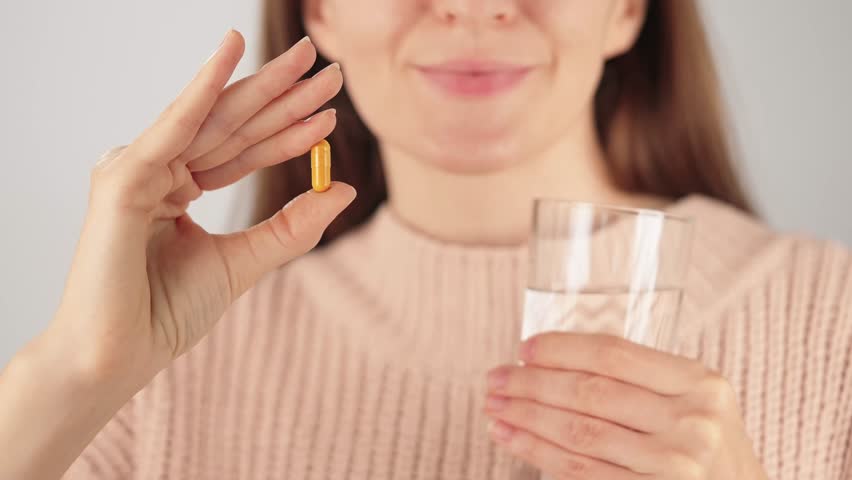 Close-up of an unrecognizable woman smiling while holding a yellow capsule and a glass of water. Concept of herbal supplements, vitamins and daily wellness routine.