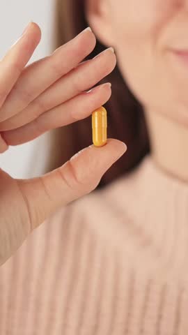 Vertical close-up of a hand holding a yellow capsule against a neutral background. Concept of herbal supplements, vitamins and daily healthcare routine.
