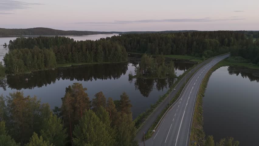 Aerial view of a Finnish road at sunset winding between two lake shores in spring, where golden light reflects on the calm water creating a peaceful and poetic landscape