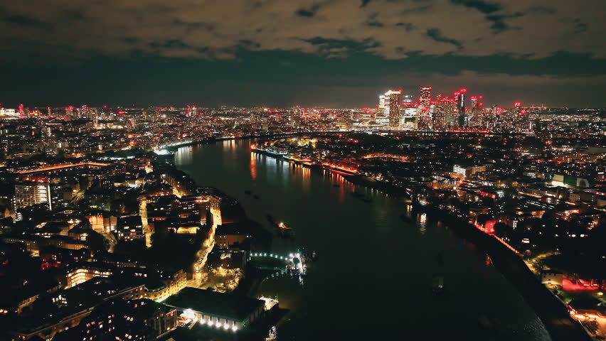 England, London: Canary Wharf skyscrapers reflecting on River Thames at night, aerial view of London urban cityscape with colorful illuminated buildings under dark cloudy sky. Drone flight footage