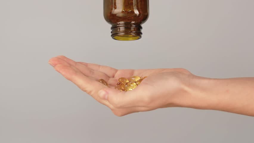 Close-up of a hand filled with golden softgel capsules being poured from a brown bottle. Concept of vitamins, fish oil, omega 3 supplements, healthcare and daily wellness routine.