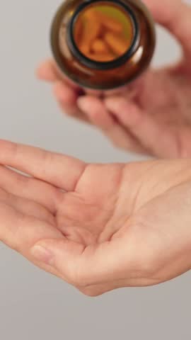 Vertical close-up of a hand full of yellow capsules being poured from a bottle. Concept of vitamins, herbal supplements, turmeric curcumin, medication and daily healthcare routine.