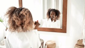 Young African American woman applying foundation with brush on her face in the bathroom mirror. Beautiful black woman in bathrobe doing morning makeup routine. Skincare, beauty, self care, and morning - Powered by Shutterstock - Get 15% off with code: PIKWIZARD15