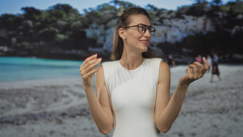 Woman in white sleeveless top smiling with hands open and gesturing at beach shore, wearing glasses and pearl earrings; joy.