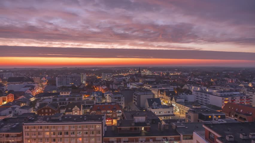 Sunrise paints the city in warm colors as night turns into day. Lights fade while the skyline awakens under glowing clouds. Timelapse of the rising sun over Sylt.