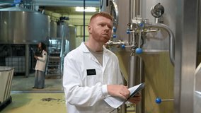 Male worker in a white coat inspecting stainless steel tanks in a brewery, checking data on a clipboard while a female colleague works in the background of the industrial beer factory - Powered by Shutterstock - Get 15% off with code: PIKWIZARD15