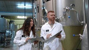 Male and female brewery professionals in white lab coats discussing production data from clipboards while inspecting large stainless steel fermentation tanks in a modern craft beer factory - Powered by Shutterstock - Get 15% off with code: PIKWIZARD15
