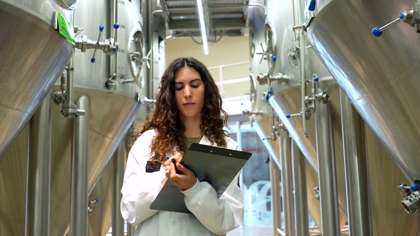 Focused female brewmaster in a white lab coat inspecting large stainless steel fermentation tanks, diligently taking notes on a clipboard during a quality control check in a modern beer factory