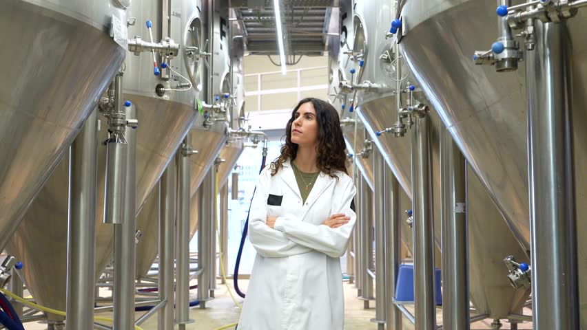 Professional brewmaster in a white lab coat proudly standing with arms crossed in a modern craft beer factory, confidently checking the stainless steel fermentation tanks and equipment