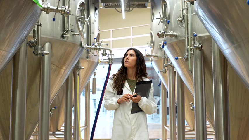 Confident brewmaster in a white lab coat walking through the brewery, diligently inspecting stainless steel tanks and taking notes on a clipboard to ensure quality control of the beer