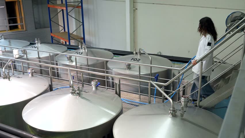Female quality control expert in a white lab coat walking on a catwalk, inspecting large stainless steel fermentation tanks and equipment during the brewing process inside a modern beer factory