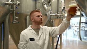 Young male brewer in a white coat pouring a glass of fresh craft beer from a fermentation tank and checking its quality, color, and aroma before smiling with satisfaction - Powered by Shutterstock - Get 15% off with code: PIKWIZARD15