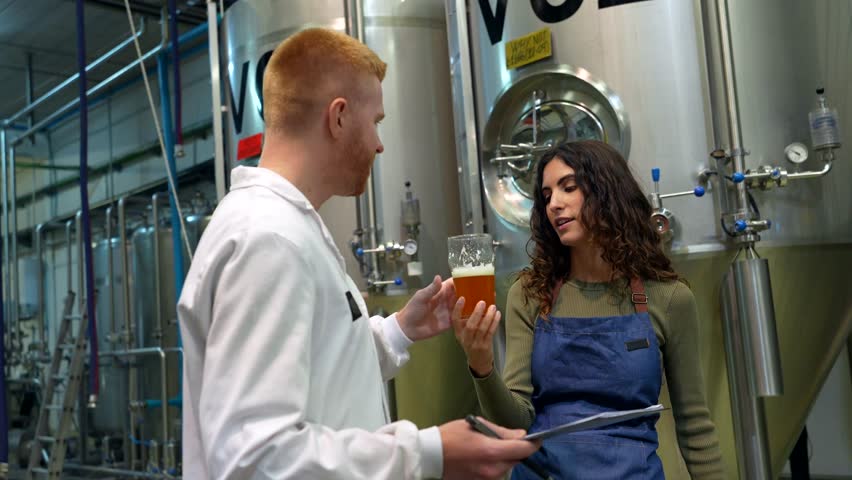 Two skilled brewery workers, a brewmaster and an assistant, analyzing the quality, color, and taste of craft beer next to large steel fermentation tanks inside an industrial production facility