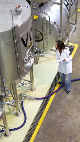 High angle view of a female quality control inspector in a white lab coat monitoring the brewing process, checking stainless steel fermentation tanks and taking notes in a modern craft beer factory