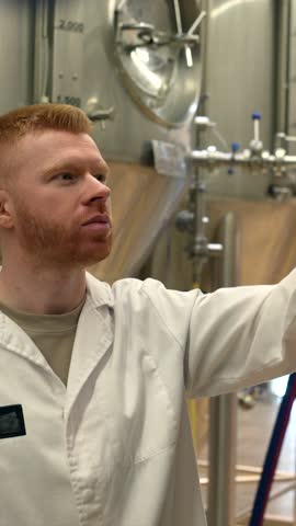 Knowledgeable brewmaster with red hair and a beard wearing a white lab coat smelling and tasting a glass of freshly brewed beer, evaluating its quality in front of large steel fermentation tanks