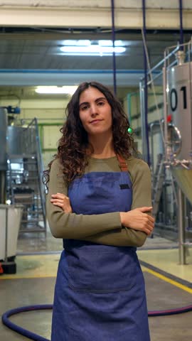 Portrait of a proud young female brewer with curly hair wearing an apron, standing with her arms crossed inside a craft brewery while looking at the camera with a confident smile