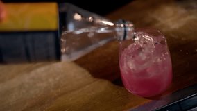 Close-up of sparkling water being poured into a pink iced cocktail in a clear rocks glass on a wooden bar, followed by the finished drink resting with ice cubes and subtle bubbles - Powered by Shutterstock - Get 15% off with code: PIKWIZARD15