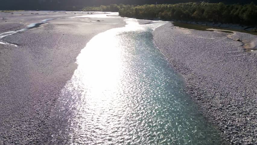 Dolly drone shot of the Tagliamento river, highlighting sun reflections on water, surrounding terrain and ending with a reveal of distant mountain peaks in daylight