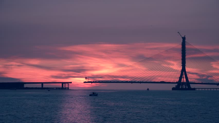 Cloudy sunset with yellow orange pink sky over Tamkang Bridge silhouette and passing boats at Tamsui Taiwan during early autumn dusk.