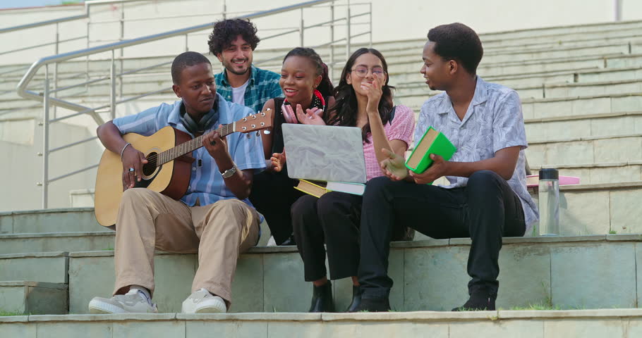 African young adult gen z man woman sitting on stair hold guitar play singing song together have fun joy outdoor place. Indian Happy smiling girl boy spend break time enjoy new day outside stairway