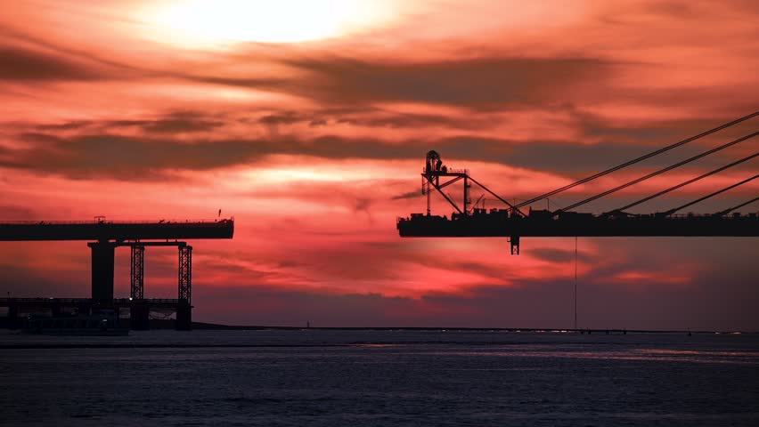 Vibrant sunset with sun partially obscured by clouds over the Tamkang Bridge construction site in Tamsui New Taipei Taiwan. Boat passes under the red and orange sky.