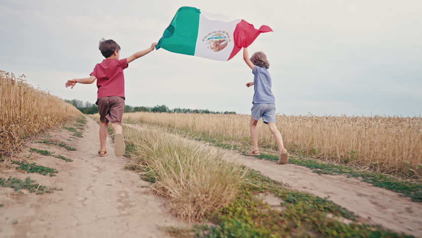 Two cheerful boys running with Mexican flag along rural road, sunny countryside