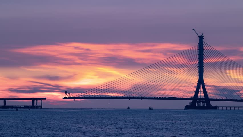 Vivid pink and purple sunset over the impressive Tamkang Bridge cable-stayed bridge construction in Tamsui New Taipei Taiwan. Boats pass by at dusk.