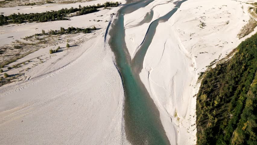 High-altitude dolly drone shot over the Tagliamento river, capturing sun reflections on water, surrounding terrain, and ending with a reveal of a road bridge and distant mountain peaks