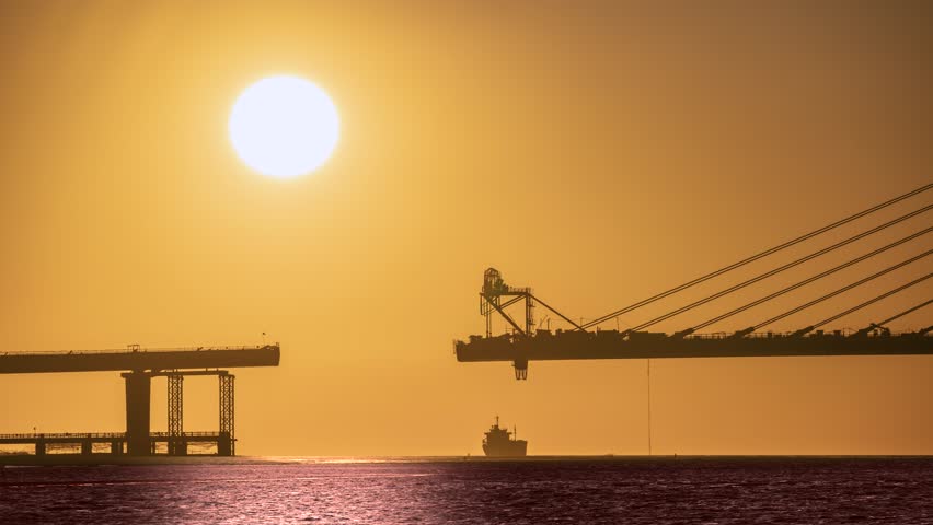 Golden hour sunset over the Tamkang Bridge construction in Tamsui New Taipei Taiwan. A ship passes under the huge setting sun, creating a dramatic silhouette scene.