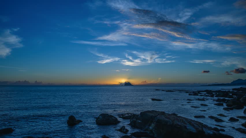 Stunning sunrise over the Xiao Jilong Islet on the horizon near Wanli New Taipei Taiwan. Golden light reflects on the calm ocean and rocky coast. Vibrant blue and orange sky.