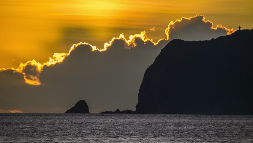 Close-up of the sun rising over the ocean near Wanli New Taipei Taiwan. Golden sky and clouds silhouette the coastal mountain and Xiao Jilong Islet in a dramatic scene.