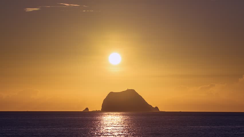 Close-up of the sun rising over the ocean near Wanli New Taipei Taiwan. Golden sky and clouds silhouette the coastal mountain and Xiao Jilong Islet in a dramatic scene.