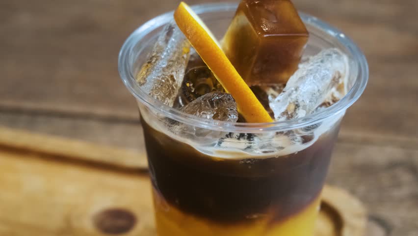 Layered drink of espresso coffee and iced orange juice in a glass with orange slice served in plastic cup for take away on wooden tray in cafe. Bumble coffee tropical summer refreshing beverage