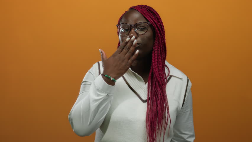 Woman smiling with a playful gesture against an orange wall, showcasing her vibrant red braids and stylish glasses in a cheerful isolated setting.