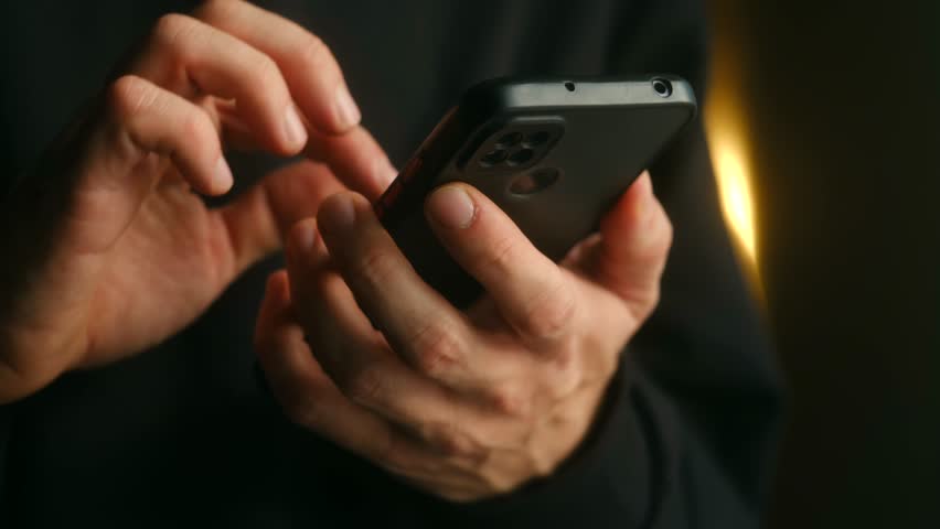 Man hands holding black smartphone, typing on touchscreen with his thumbs, scrolling through social media feed or browsing online in dark room with warm light. Person hands typing on smartphone