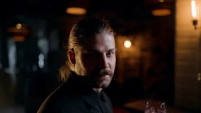 Moody bar scene showing a mustached man with tied-back hair moving from a direct gaze to savoring a pink cocktail beneath warm Edison bulbs in a dim, intimate lounge setting. - Powered by Shutterstock - Get 15% off with code: PIKWIZARD15