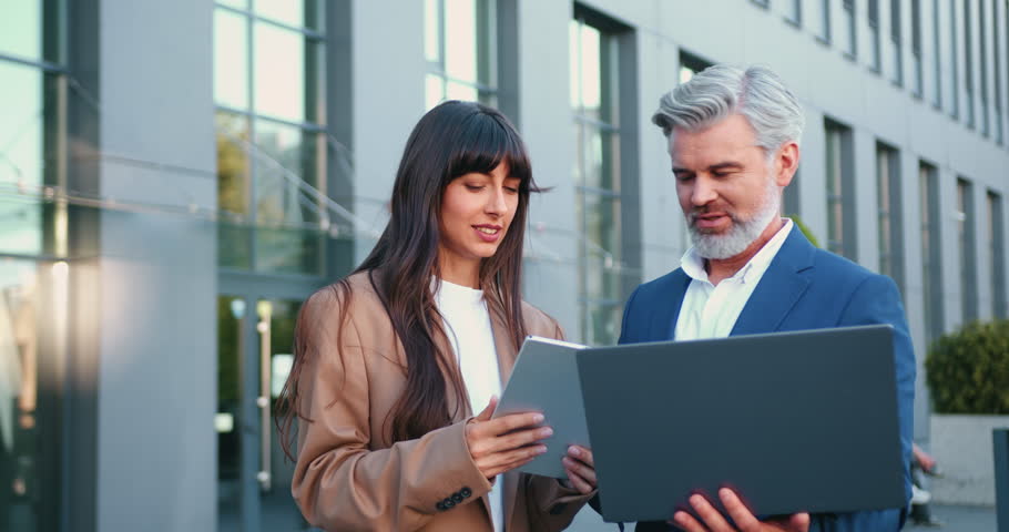 Close-up of mature businessman with laptop and stylish young businesswoman with tablet standing outside modern office building, collaborating on project and discussing business strategy