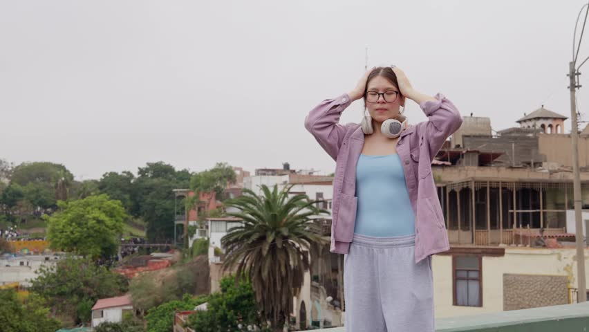 Young woman with headphones enjoying music and dancing freely on a rooftop. Expressive and energetic performance against an urban cityscape