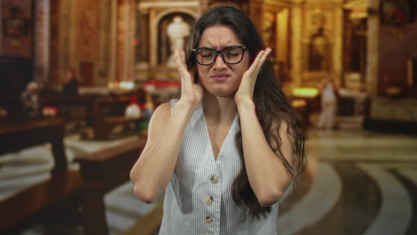 Young hispanic woman pressing ears to head with pained expression in ornate church building interior under warm ambient light; discomfort.