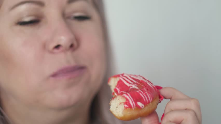 Close-up of a middle-aged Caucasian woman enjoying a sweet round donut with icing, woman eating dessert, slow motion
